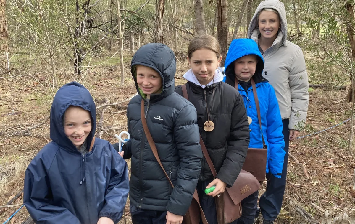 A group of children and an adult are standing in a wooded area.