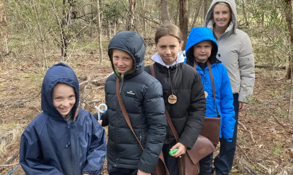 A group of children and an adult are standing in a wooded area.
